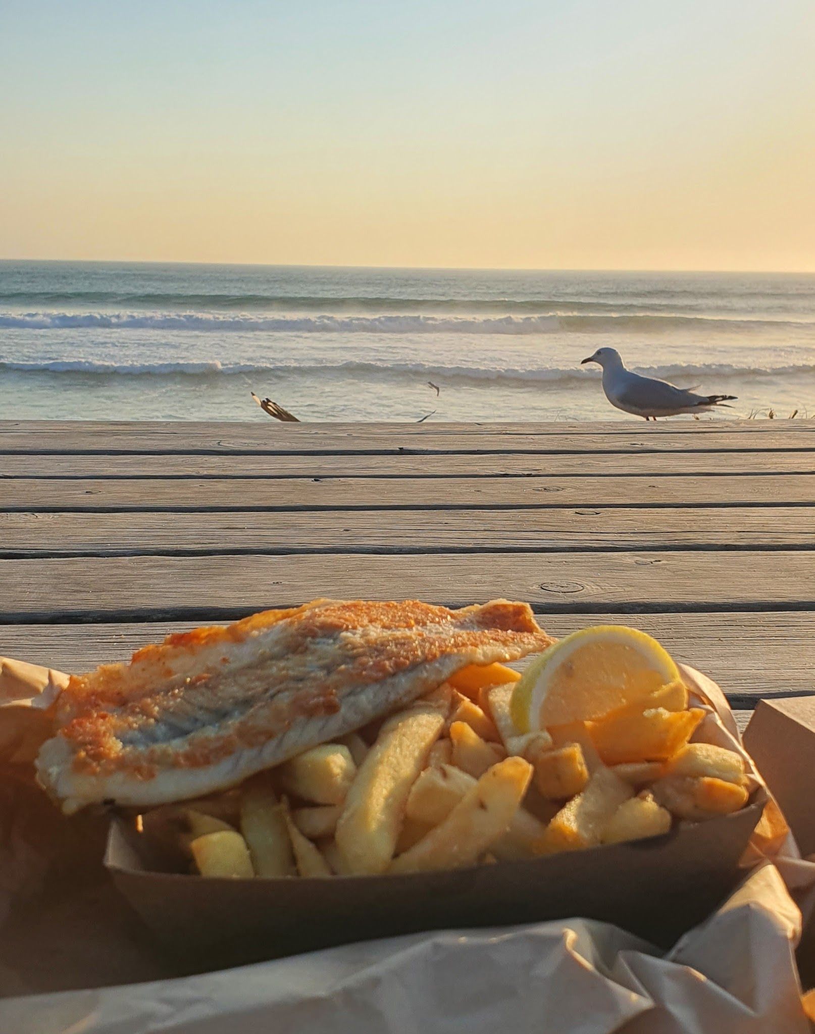 Fish and chips on a beach deck at golden hour with ocean waves and a seagull in the background